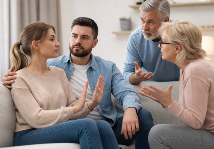 a distressed couple on a couch with parents, one gesturing stop, representing setting boundaries with in-laws.