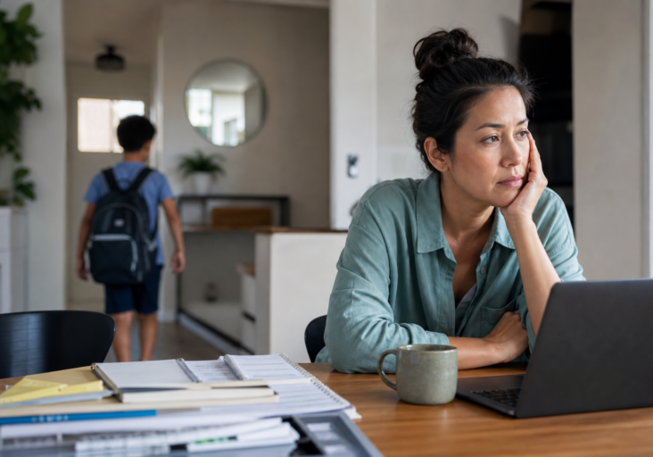 a pensive woman at a laptop experiencing parent back-to-school anxiety as her child walks away with a backpack.
